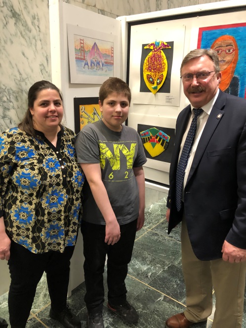 On March 6, 2019, Assemblyman Brian Miller, Joshua Ellis, and Josh’s mom, Vickie Ellis, pose next to Josh’s collage, “Relief Mask,” now on display in the Legislative Office Buildin